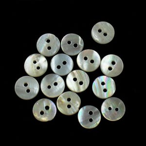 May include: A collection of 14 white mother of pearl buttons with two holes each. The buttons are arranged in a random pattern on a black background.