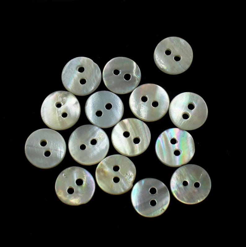 May include: A collection of 14 white mother of pearl buttons with two holes each. The buttons are arranged in a random pattern on a black background.
