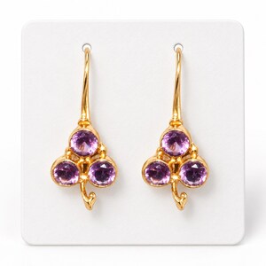 May include: A pair of gold drop earrings featuring a clover-shaped design. Each earring is set with three round, faceted purple amethyst gemstones and includes a curved gold wire hook for attachment, creating a symmetrical floral appearance.
