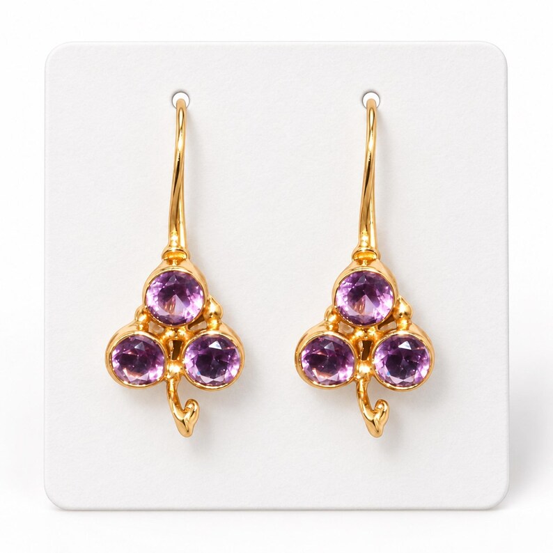 May include: A pair of gold drop earrings featuring a clover-shaped design. Each earring is set with three round, faceted purple amethyst gemstones and includes a curved gold wire hook for attachment, creating a symmetrical floral appearance.