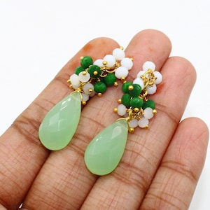 May include: A pair of gold-toned earrings featuring a cluster of green and white beads above a light green teardrop-shaped stone. The earrings are displayed against a neutral background.
