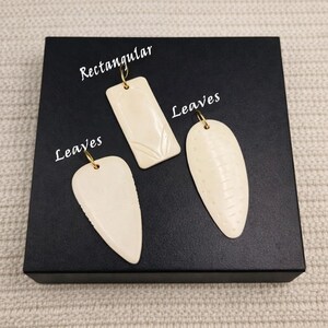 May include: Three off-white pendants with gold-colored loops. One is rectangular with a leaf design, and two are leaf-shaped. The word "Rectangular" is above the rectangular pendant, and the word "Leaves" is above the leaf-shaped pendants.