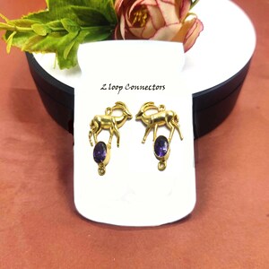 May include: Two gold-colored deer-shaped pendants with oval, purple gemstones. The pendants are displayed on a white card with the text "Z loop Connectors". The background includes a flower and greenery.