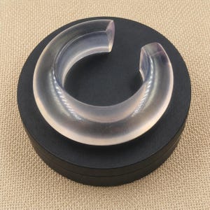 May include: A clear, translucent, C-shaped cuff bracelet with a smooth, rounded surface. The bracelet is positioned on a circular, matte black display stand. The design features an open gap, creating a continuous, curved, semi-circular form.
