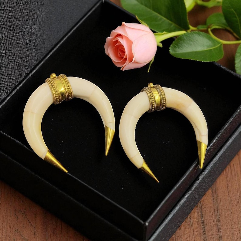 May include: A pair of ivory-coloured, crescent-shaped earrings with gold-coloured accents. The earrings have a band of gold near the top and pointed gold tips. They are displayed in a black box with a pink rose in the background.