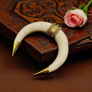 May include: A curved, off-white horn-shaped pendant with gold-coloured tips and a decorative gold band. The pendant is set against a dark wooden surface, with a pink rose in the background. The pendant is likely a piece of jewellery.