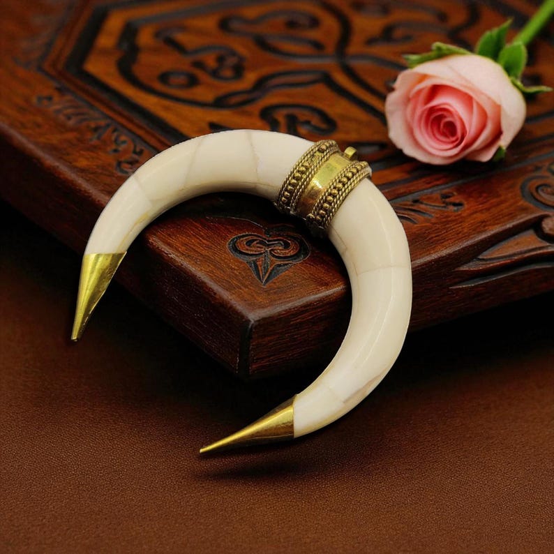 May include: A curved, off-white horn-shaped pendant with gold-coloured tips and a decorative gold band. The pendant is set against a dark wooden surface, with a pink rose in the background. The pendant is likely a piece of jewellery.