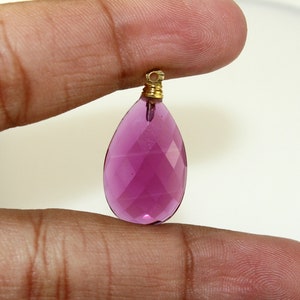 May include: A faceted, teardrop-shaped pendant in a vibrant magenta hue. The gemstone is suspended from a gold-colored metal bail. The pendant is held between two fingers against a plain white background.