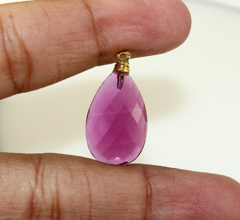 May include: A faceted, teardrop-shaped pendant in a vibrant magenta hue. The gemstone is suspended from a gold-colored metal bail. The pendant is held between two fingers against a plain white background.
