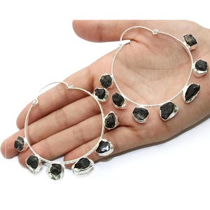 May include: Silver hoop earrings with black gemstone accents. The earrings are large and have a modern, minimalist design.