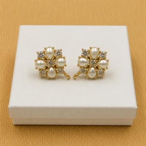 May include: A pair of gold-toned earrings, each featuring a floral design. Each earring has six pear-shaped faux pearls and six clear, round-cut crystals. The earrings are presented on a white box against a gold background.