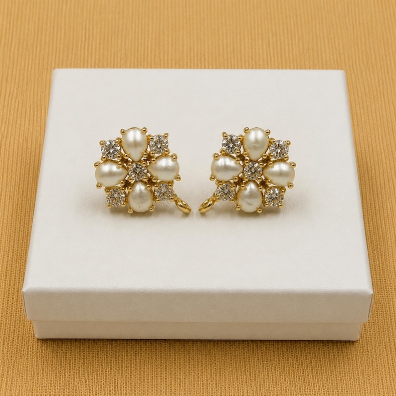 May include: A pair of gold-toned earrings, each featuring a floral design. Each earring has six pear-shaped faux pearls and six clear, round-cut crystals. The earrings are presented on a white box against a gold background.