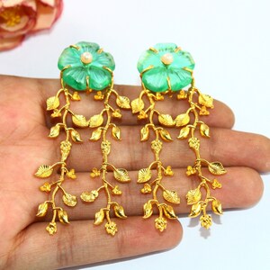 Puede incluir: Un par de pendientes de oro con piedras verdes en forma de flor y un acento de perla. Los pendientes presentan una delicada cadena de hojas doradas.