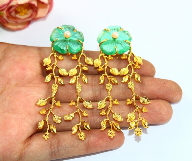 Puede incluir: Un par de pendientes de oro con piedras verdes en forma de flor y un acento de perla. Los pendientes presentan una delicada cadena de hojas doradas.