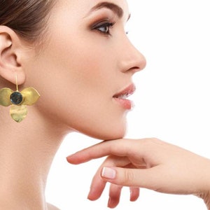 May include: Gold-coloured earrings with a floral design. Each earring features three petal-shaped elements and a central, dark-coloured stone. The earrings are suspended from a thin wire, showcasing a unique, nature-inspired aesthetic.