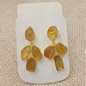 May include: A pair of gold-toned earrings featuring irregularly shaped, amber-colored stones. The stones are set in gold-colored metal and arranged in a cascading design. The earrings are displayed on a white card.