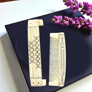 May include: Two antique ivory-colored combs. One is a folding comb with a decorative crisscross pattern. The other is a straight comb with a decorative border. Both combs have black etched details. A sprig of purple flowers is in the background.