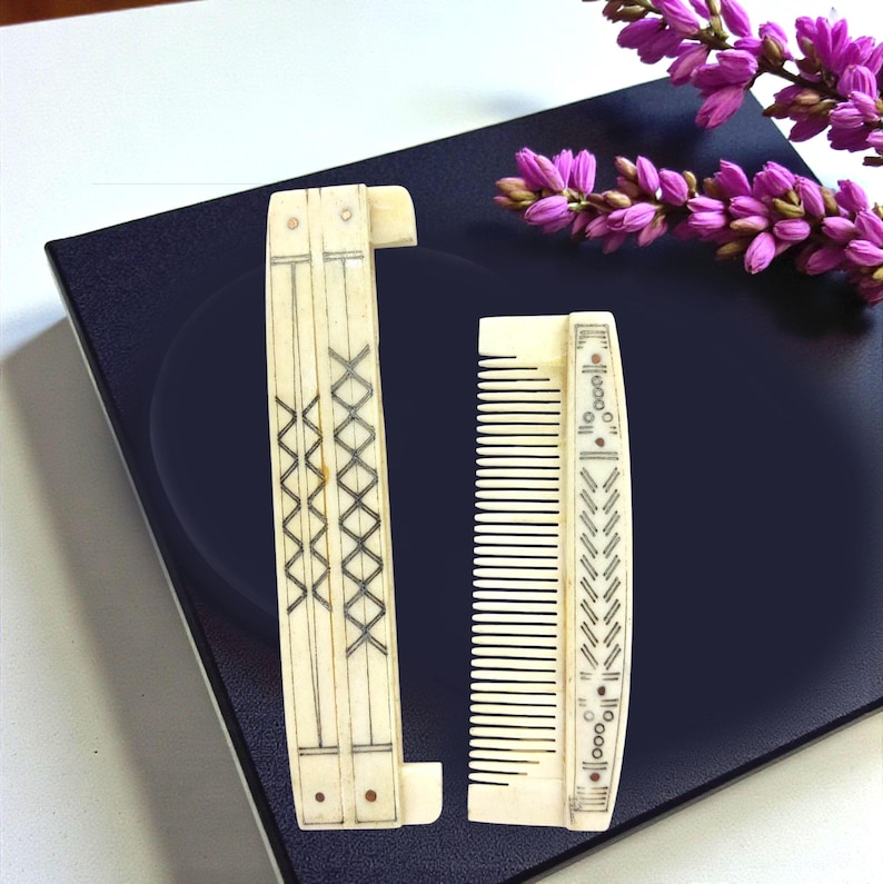 May include: Two antique ivory-colored combs. One is a folding comb with a decorative crisscross pattern. The other is a straight comb with a decorative border. Both combs have black etched details. A sprig of purple flowers is in the background.