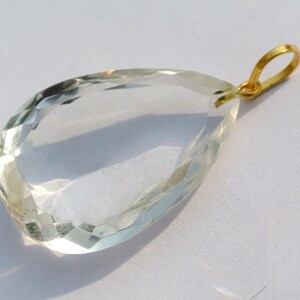 May include: A clear, faceted teardrop-shaped pendant with a gold-colored bail. The gemstone is transparent with a subtle green hue, catching the light to create a sparkling effect. The pendant is likely a piece of jewelry.