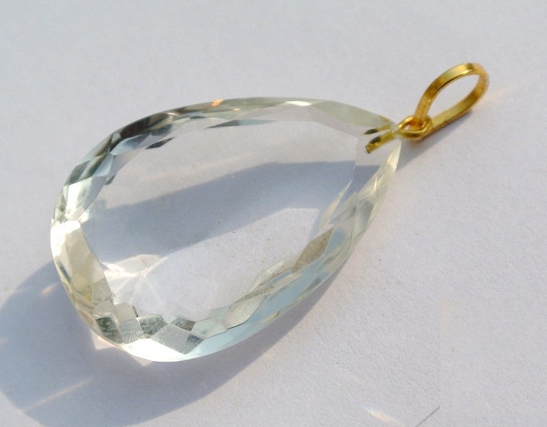 May include: A clear, faceted teardrop-shaped pendant with a gold-colored bail. The gemstone is transparent with a subtle green hue, catching the light to create a sparkling effect. The pendant is likely a piece of jewelry.