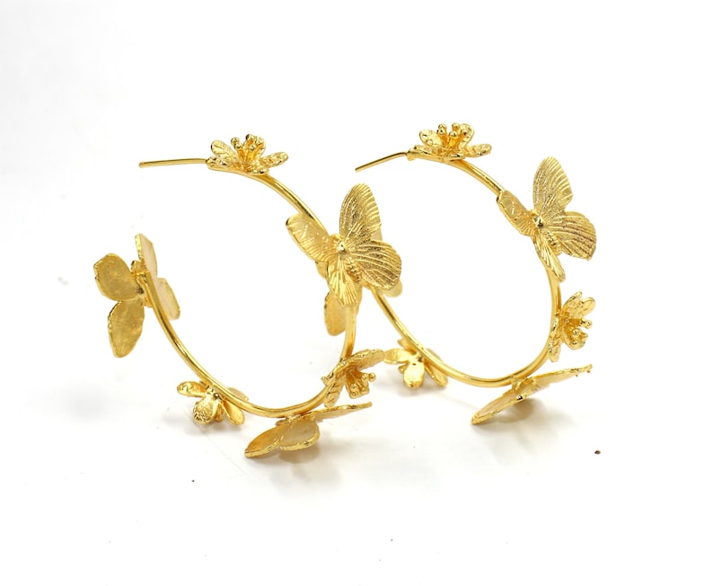 Puede incluir: Aretes de aro dorados con un delicado dise&ntilde;o de mariposa y flores. Los aretes est&aacute;n hechos de metal y tienen un acabado pulido.