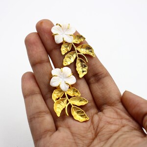 May include: A pair of gold-colored earrings featuring white flower-shaped accents and gold-colored leaf designs. The earrings are designed to resemble a floral arrangement, with the flowers at the top and the leaves extending downwards.