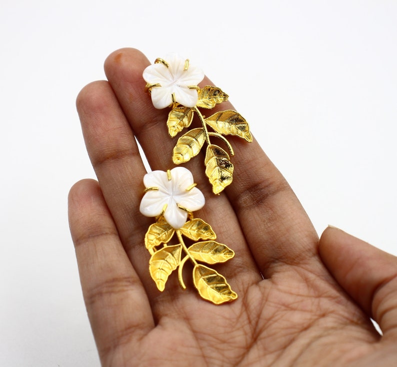 May include: A pair of gold-colored earrings featuring white flower-shaped accents and gold-colored leaf designs. The earrings are designed to resemble a floral arrangement, with the flowers at the top and the leaves extending downwards.