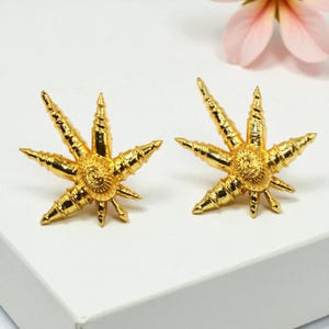 May include: A pair of gold-colored starburst earrings. Each earring features a central circular design with radiating points, creating a star-like shape. The earrings have a textured surface and a bright, shiny finish.
