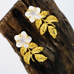 May include: A pair of floral earrings featuring white flower-shaped pendants with gold-colored accents. The earrings have gold-colored leaf and stem designs. The earrings are displayed on a dark brown wooden surface.