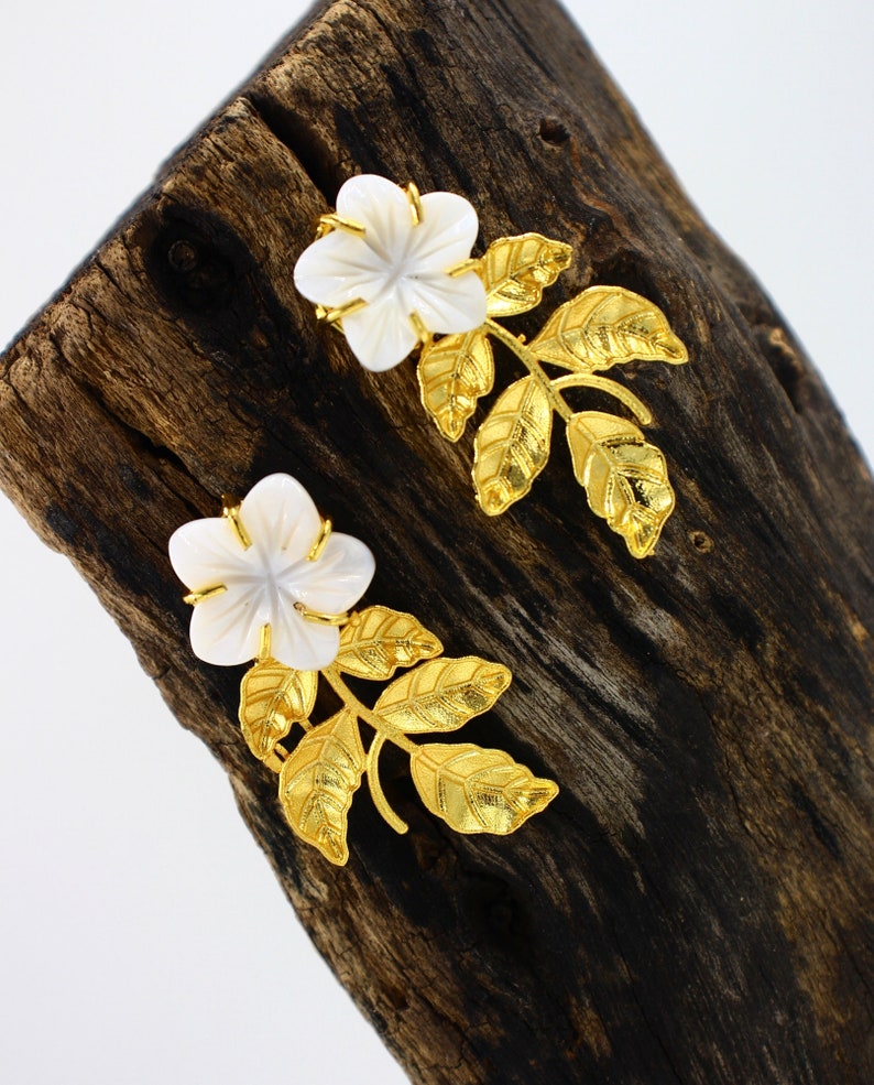 May include: A pair of floral earrings featuring white flower-shaped pendants with gold-colored accents. The earrings have gold-colored leaf and stem designs. The earrings are displayed on a dark brown wooden surface.