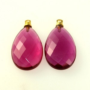 May include: Two faceted, teardrop-shaped pendants. The pendants are a deep magenta color with gold-colored caps. The facets create a sparkling effect, and the overall design is elegant and eye-catching. These pendants are suitable for jewelry making.