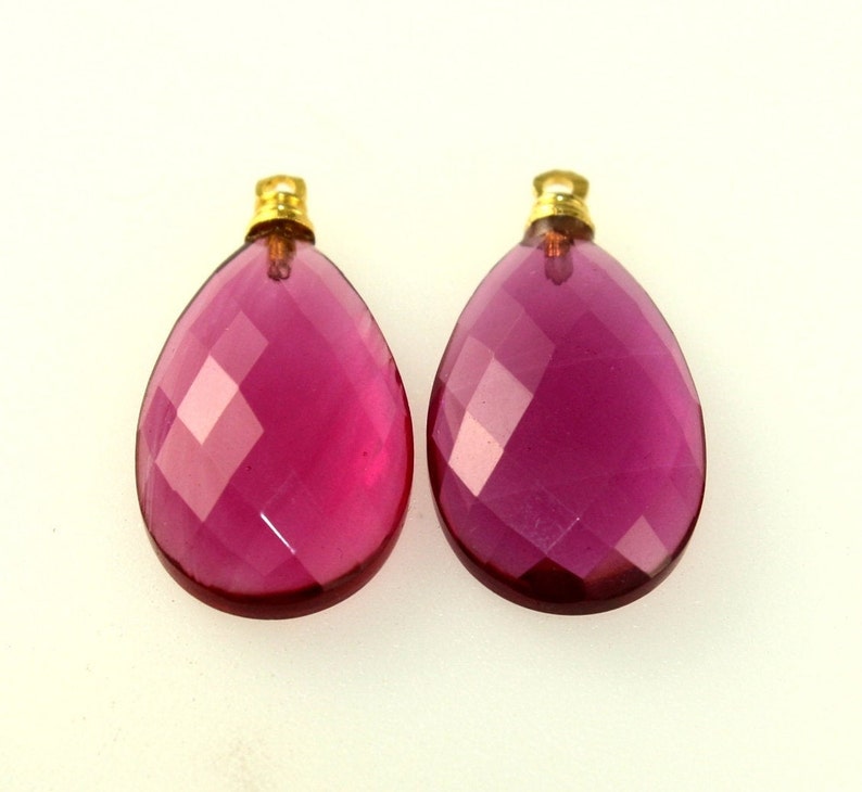 May include: Two faceted, teardrop-shaped pendants. The pendants are a deep magenta color with gold-colored caps. The facets create a sparkling effect, and the overall design is elegant and eye-catching. These pendants are suitable for jewelry making.