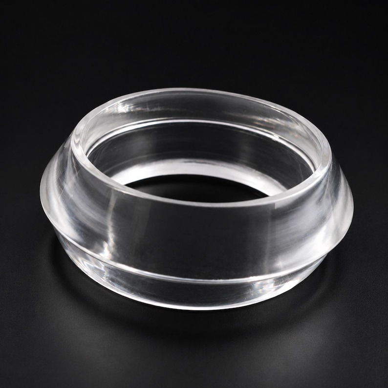 May include: A clear, transparent, circular bangle bracelet with a smooth, polished surface. The bracelet features a wide, slightly tapered band design with a rounded outer edge and a hollow center, creating a simple, uniform, and reflective appearance.