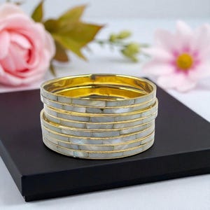 Può includere: Una pila di braccialetti dorati con intarsio in madreperla. I braccialetti sono impilati su una scatola nera, con fiori rosa sullo sfondo. I braccialetti hanno una superficie lucida e riflettente.