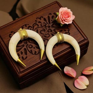 May include: Two ivory-coloured, crescent-shaped pendants with gold-coloured metal accents. The pendants are displayed on a dark wooden box with floral carvings, alongside a pink rose and petals. The pendants have a gold band and pointed tips.