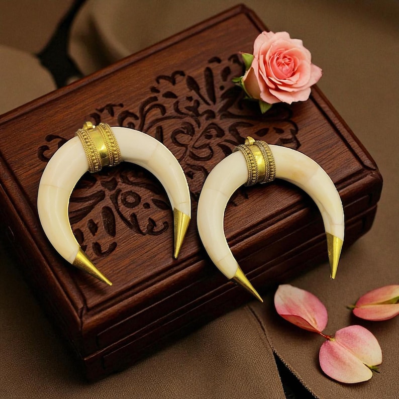 May include: Two ivory-coloured, crescent-shaped pendants with gold-coloured metal accents. The pendants are displayed on a dark wooden box with floral carvings, alongside a pink rose and petals. The pendants have a gold band and pointed tips.