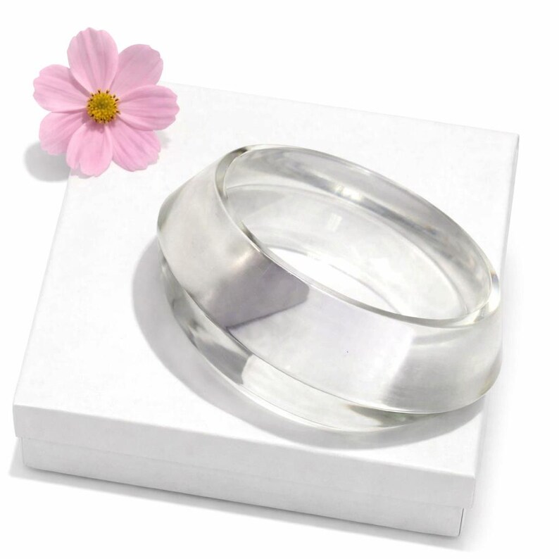 May include: Clear, transparent bangle bracelet with a rounded design. The bracelet is resting on a white surface, next to a pink flower with a yellow center. The bangle is a simple, elegant accessory.