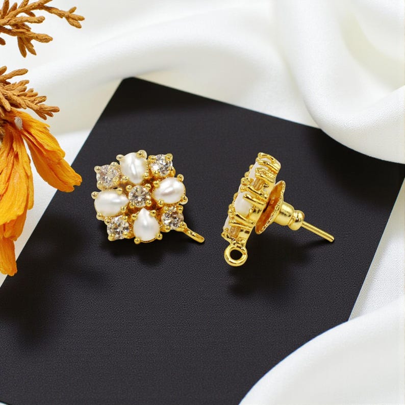 May include: A pair of gold-toned stud earrings. Each earring features a cluster of white pearls and clear crystals, arranged in a floral design. The earrings are set against a black background with a white fabric.