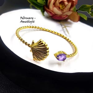 May include: Gold-tone bracelet with a twisted design, featuring a seashell charm and a purple amethyst gemstone. "February - Amethyst" is written on the background.