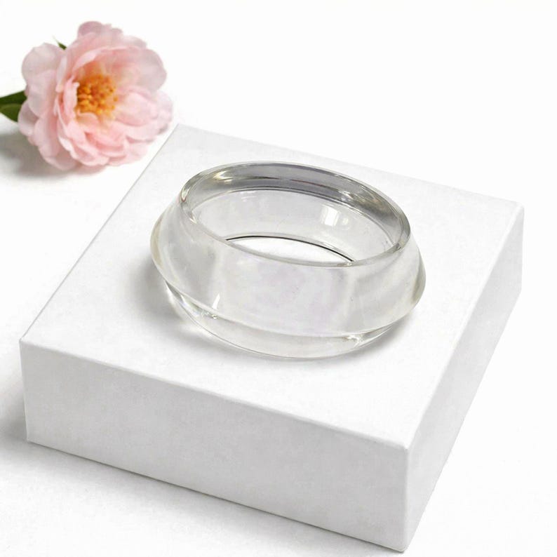 May include: A clear, circular glass bracelet sits on a white box, with a pink flower in the background. The bracelet has a rounded, layered design. The overall aesthetic is minimalist and elegant.