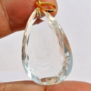 May include: A clear, teardrop-shaped pendant with a faceted surface. The gemstone is suspended from a small, gold-colored bail. The pendant is held up by a finger.