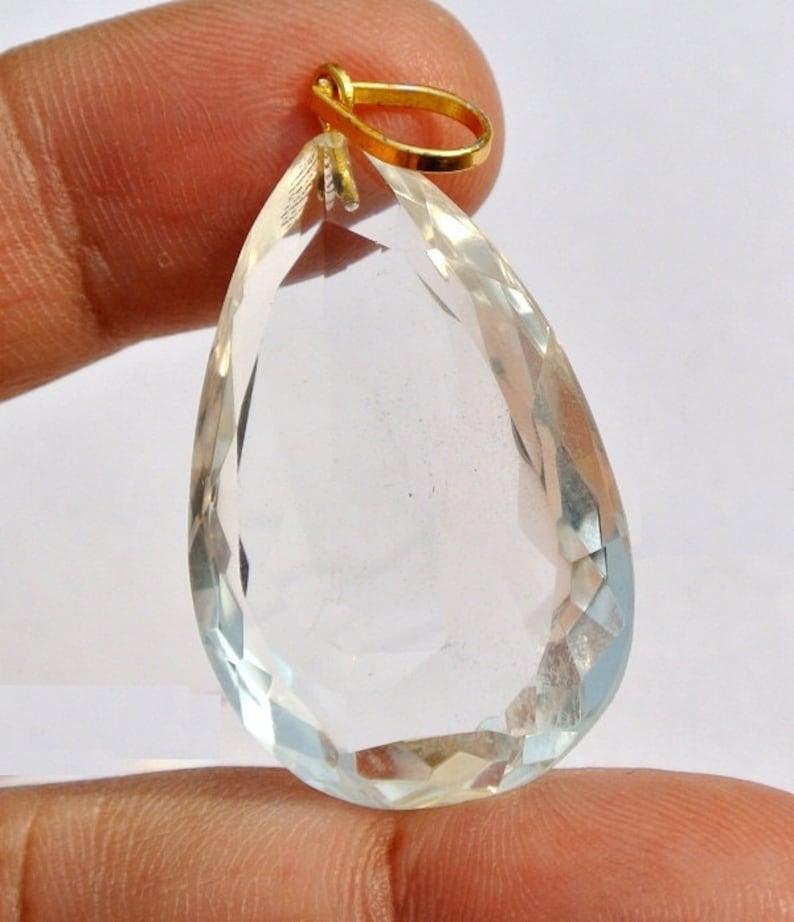 May include: A clear, teardrop-shaped pendant with a faceted surface. The gemstone is suspended from a small, gold-colored bail. The pendant is held up by a finger.