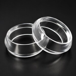 May include: Two clear, transparent rings with a smooth, polished finish. Each ring features a distinct, raised central band that encircles the circumference, creating a layered, geometric profile with a glossy, reflective surface texture.