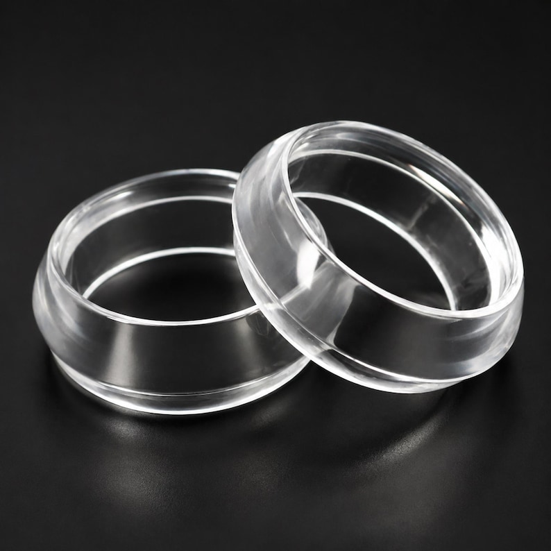May include: Two clear, transparent rings with a smooth, polished finish. Each ring features a distinct, raised central band that encircles the circumference, creating a layered, geometric profile with a glossy, reflective surface texture.