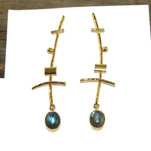 Puede incluir: Pendientes de oro con un diseño geométrico y una piedra ovalada azul. Los pendientes están hechos de alambre de oro y tienen un acabado texturizado.