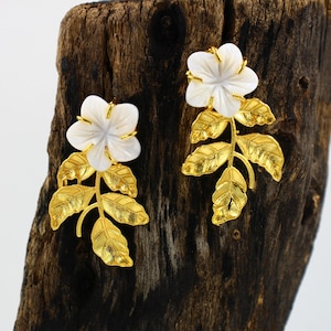 May include: A pair of floral earrings. Each earring features a white flower with a gold-colored center, set atop gold-colored leaves and stems. The earrings are designed to resemble a flower and leaf arrangement.