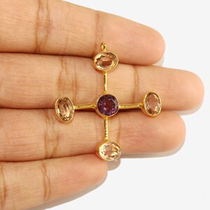 Puede incluir: Colgante dorado con dise&ntilde;o de cruz. El centro presenta una gema redonda de color p&uacute;rpura, rodeada por cuatro gemas ovaladas de color claro. El colgante se sostiene en una mano sobre un fondo blanco.