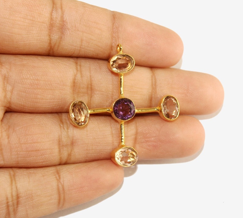 Puede incluir: Colgante dorado con dise&ntilde;o de cruz. El centro presenta una gema redonda de color p&uacute;rpura, rodeada por cuatro gemas ovaladas de color claro. El colgante se sostiene en una mano sobre un fondo blanco.
