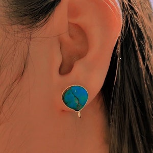 May include: A close-up of a person's ear with a turquoise gemstone earring. The earring has a silver setting and a small, dangling charm.