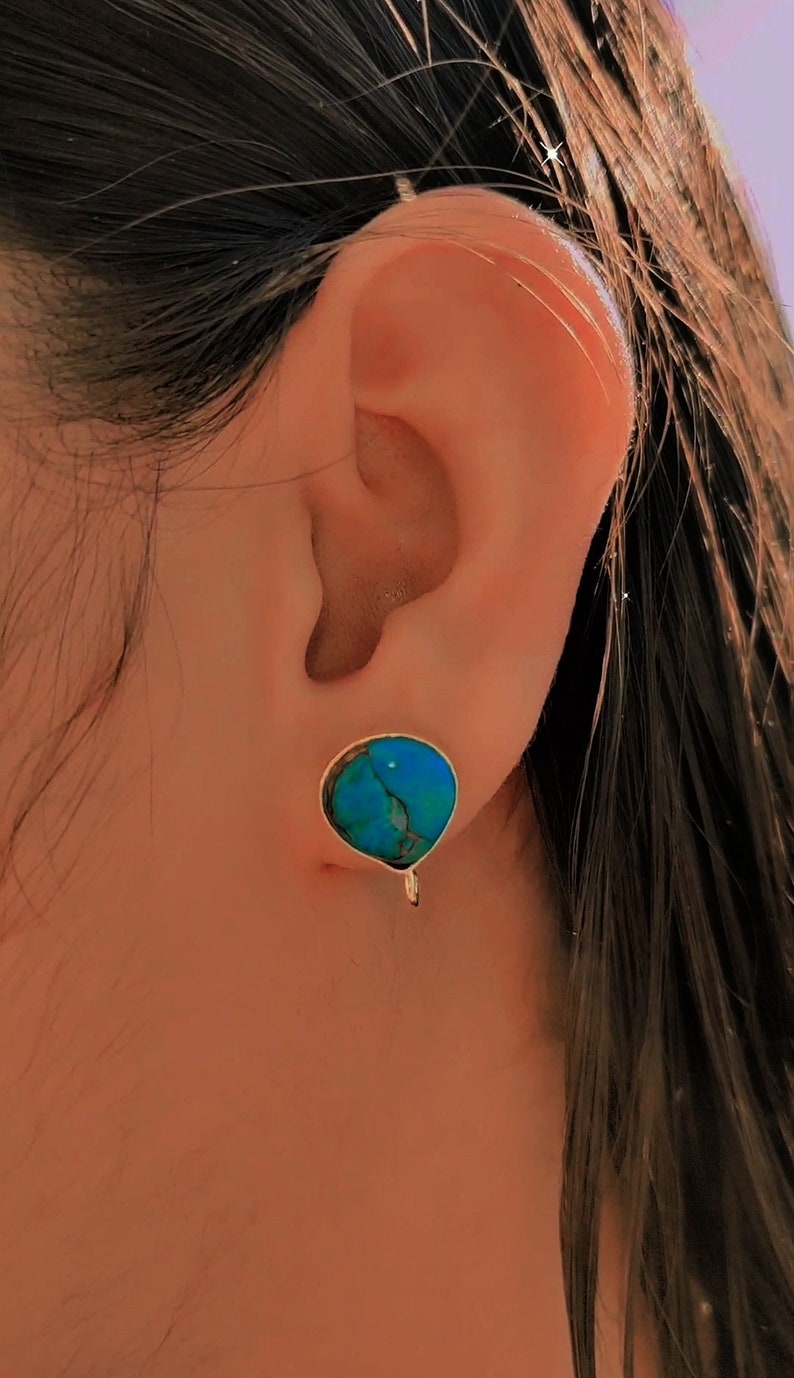 May include: A close-up of a person's ear with a turquoise gemstone earring. The earring has a silver setting and a small, dangling charm.